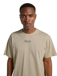 Mand med kort hår og lille ring-ørestik iført beige t-shirt med teksten "HALO" trykt foran, vendt mod fronten mod hvid baggrund.