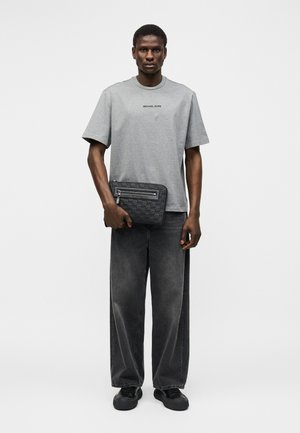 Hombre con camiseta gris de Michael Kors, vaqueros negros holgados, zapatillas negras, sosteniendo un bolso de mano negro con cremallera de Michael Kors.