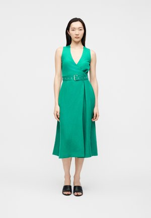 Kvinde stående iført en ærmeløs grøn midikjole med V-hals og bælte i taljen, kombineret med sorte åbne mule-sandaler.