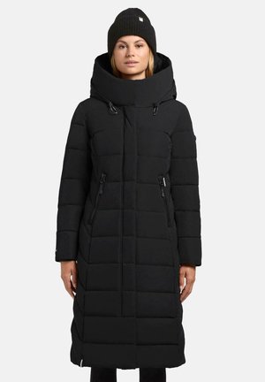 Vrouw in een lange zwarte gewatteerde winterjas met een hoge kraag en ritszakken, gecombineerd met een zwarte gebreide beanie.