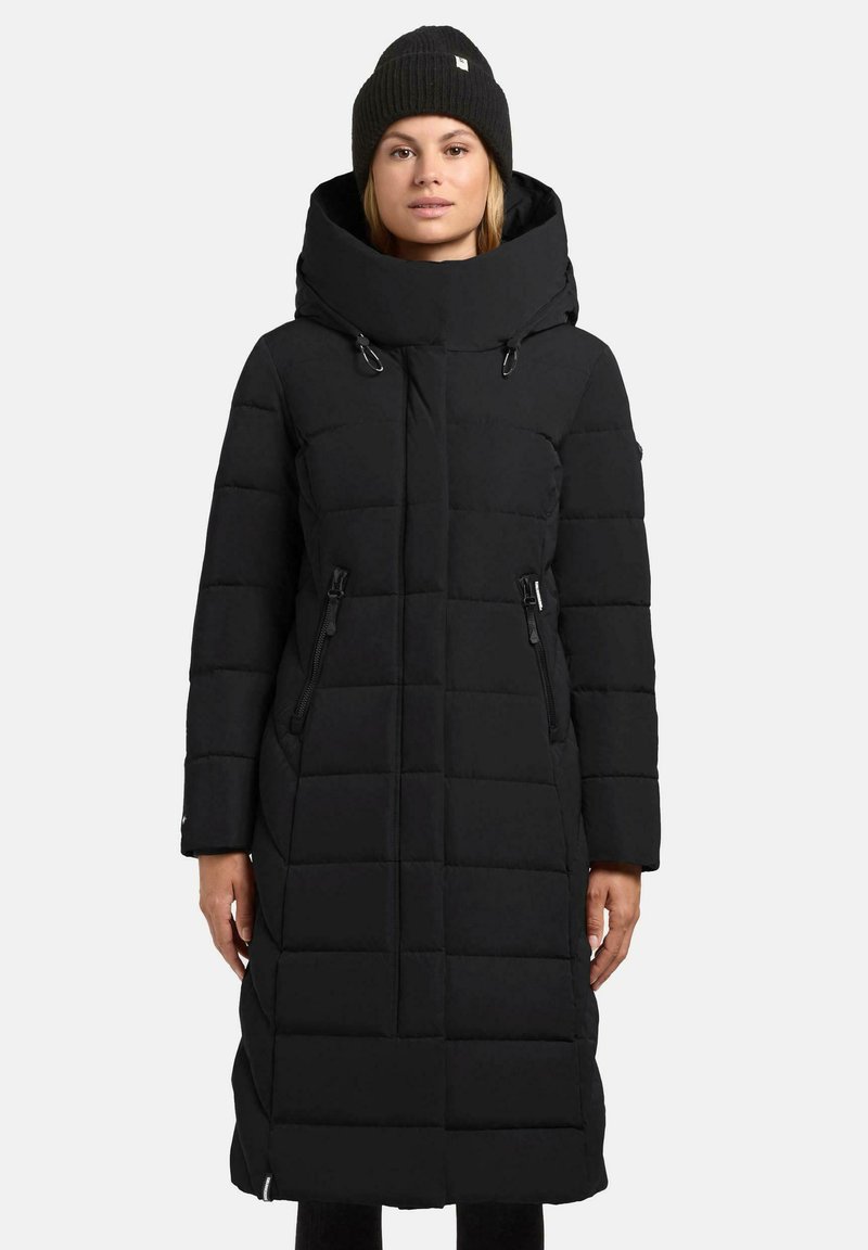Vrouw in een lange zwarte gewatteerde winterjas met een hoge kraag en ritszakken, gecombineerd met een zwarte gebreide beanie.