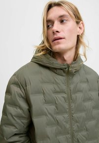 Mladý člověk s dlouhými blond vlasy, oblečený v olivově zelené prošívané bundě s kapucí, dívající se stranou na světlém jednobarevném pozadí.