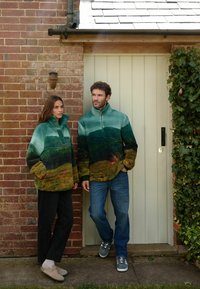 Des pulls en polaire avec un design de paysage montagneux dans des nuances de vert et de marron ; col zippé, manches larges ; associés à un denim et des chaussures aux teintes neutres.