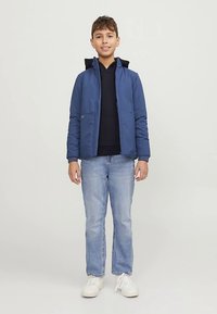 Modrá prešívaná bunda s kapucňou, nosená cez tmavú mikinu s kapucňou. Ľahké modré džínsy a biele tenisky dopĺňajú outfit. Stojí na neutrálnej pozadí.