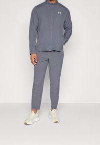 Grå idrottsjacka med hög krage, dragkedjestängning och matchande åtsittande byxor. Har en reflexlogotyp och kontrasterande sömmar.