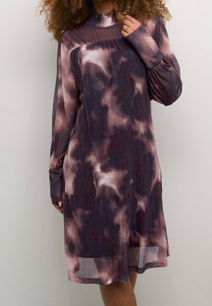 Kvinde iført en langærmet, knælange kjole med et lilla og sort tie-dye mønster, stående mod en ensfarvet lys baggrund.