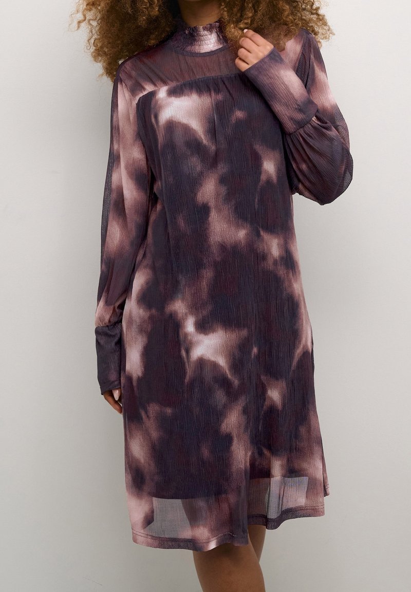 Femme portant une robe à manches longues, longueur genoux, avec un motif tie-dye violet et noir, debout devant un fond clair uni.