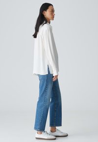 Chemise blanche à manches longues avec patte de boutonnage, associée à un jean droit bleu et des baskets blanches. Tissu lisse, coupe décontractée et fentes sur les côtés.