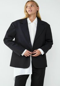 Czarna, oversize marynarka z szerokimi klapami, z zapięciem na jeden guzik, noszona na białej koszuli z zaokrąglonym dołem, w połączeniu z czarnymi spodniami.