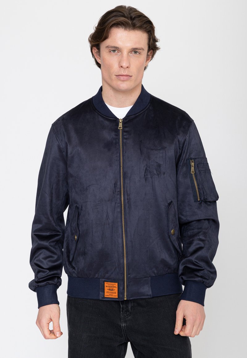 Junger Mann trägt eine marineblaue Bomberjacke mit Reißverschluss vorne und Tasche, steht vor einem schlichten weißen Hintergrund.