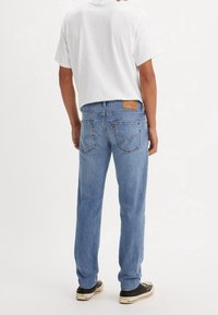 Homme portant un jean bleu clair à jambes droites, un t-shirt blanc uni à manches courtes et des baskets noires, debout de dos.
