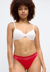 Biały stanik z cienkimi ramiączkami, gładki materiał, w zestawie z czerwonym stringami z koronkowym wykończeniem wzdłuż pasa. Modelka z kręconymi włosami pozuje frontalnie.