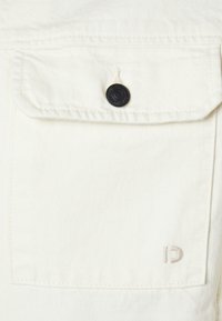 Tasca della giacca in denim bianco con chiusura a bottone nero, cuciture dettagliate e un piccolo "CD" impresso in beige chiaro nell'angolo in basso a destra.