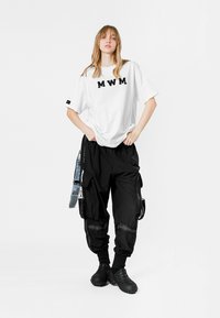 Camiseta blanca con el texto "MWM" en negro, combinada con pantalones cargo negros holgados que tienen múltiples bolsillos y correas laterales, usados con calzado negro.
