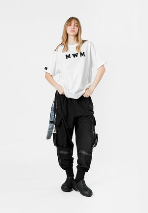 Camiseta blanca con el texto "MWM" en negro, combinada con pantalones cargo negros holgados que tienen múltiples bolsillos y correas laterales, usados con calzado negro.