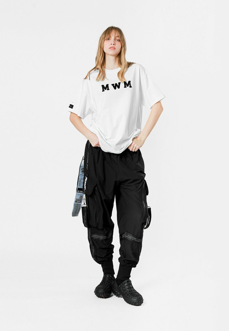 Camiseta blanca con el texto "MWM" en negro, combinada con pantalones cargo negros holgados que tienen múltiples bolsillos y correas laterales, usados con calzado negro.
