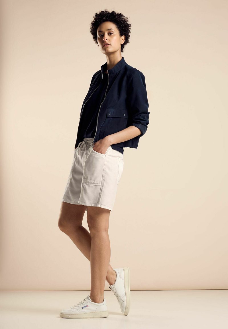 Chaqueta azul marino con cremallera, camiseta negra, pantalones cortos crema con bolsillos y zapatillas blancas. Tejidos suaves, diseño minimalista, corte relajado.