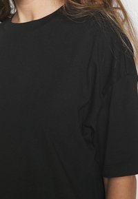 Personne portant un T-shirt noir uni à manches courtes avec de longs cheveux châtain clair visibles sur le côté droit.