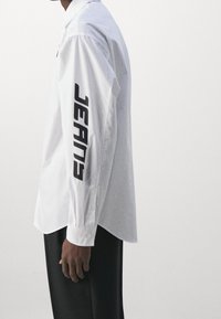 Weißes Hemd mit Knopfleiste und dem in fettem Schwarz auf dem linken Ärmel aufgedruckten Schriftzug "JEANS", mit glatter Textur und lockerem Schnitt.