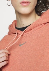 Gros plan d'une personne portant un sweat à capuche Nike corail rose, tenant le cordon de serrage, avec un petit logo Nike multicolore sur la poitrine et une boucle d'oreille argentée visible.