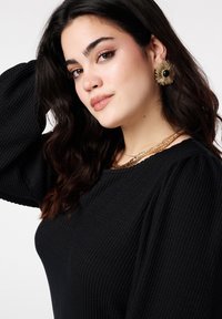 Zwarte top met textuur en opgepofte mouwen, gelaagde gouden ketting en bloemvormige oorbellen met een zwart middelpunt.