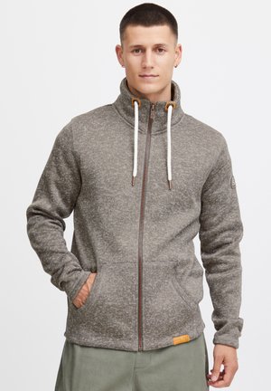 Sivi zip-up flis jakna s visokim ovratom, vezicama, dva prednja džepa i teksturiranim uzorkom. Uključuje zakrpu s logom na rukavu.
