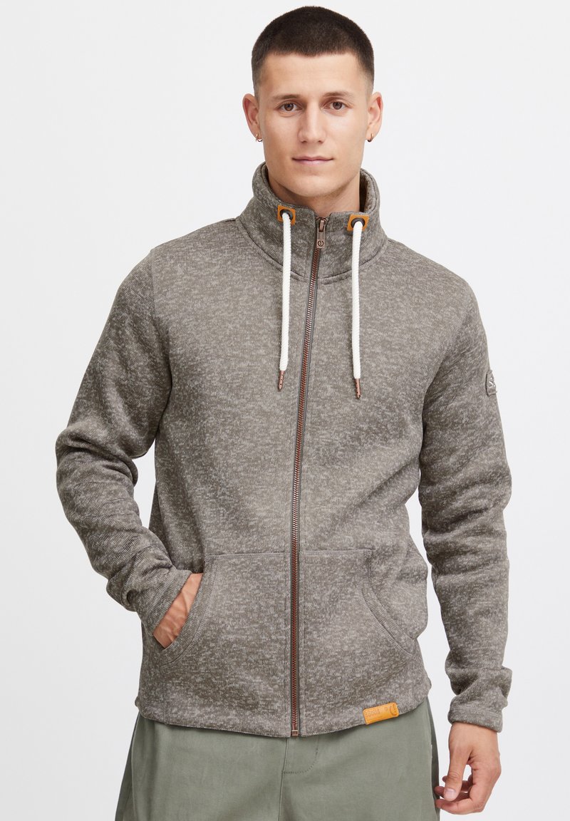 Gray zip-up fleece jacket with high collar, drawstrings, two front pockets, and a textured pattern. Includes a logo patch on the sleeve.