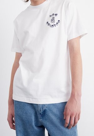 Persona che indossa una maglietta bianca New Balance con un logo della mano che fa il segno di pace sul petto, abbinata a jeans blu.