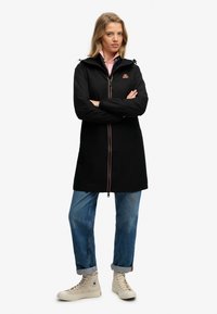 Parka impermeable negra con cierre de cremallera, puños elásticos y capucha ajustable. Presenta un logo y costuras en contraste; combinado con vaqueros azul claros y zapatillas beige.