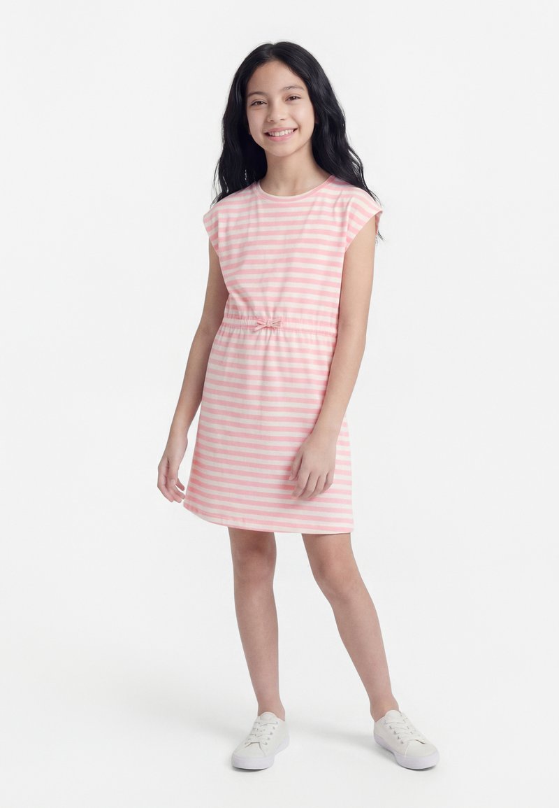 Κορίτσι με μακριά μαύρα μαλλιά που χαμογελά, φοράει φόρεμα με ροζ και λευκές ρίγες και μικρό φιόγκο, καθώς και λευκά αθλητικά παπούτσια, στέκεται μπροστά σε λευκό φόντο.