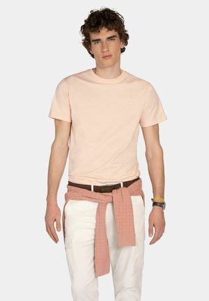 Joven con cabello rizado, vestido con camiseta rosa pálido, pantalones blancos, suéter rosa atado a la cintura, cinturón marrón y accesorios en la muñeca.