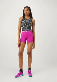 O femeie atletică stă în picioare, purtând un top fără mâneci negru-gri, pantaloni scurți Nike Pro roz aprins, sosete negre și pantofi sport roz cu talpă turcoaz.