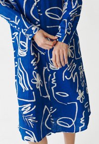 Abito in lino blu con motivi astratti bianchi, chiusura frontale con bottoni, maniche lunghe e vestibilità ampia, con orlo appena sopra le ginocchia.