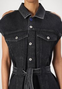 Μαύρο τζιν φόρεμα πουκαμίσα με κοντά μανίκια, μεγάλο γιακά, εμπρόσθιες τσέπες και αντίθετη λευκή ραφή. Διαθέτει ζώνη στη μέση και μπλε κουμπί.