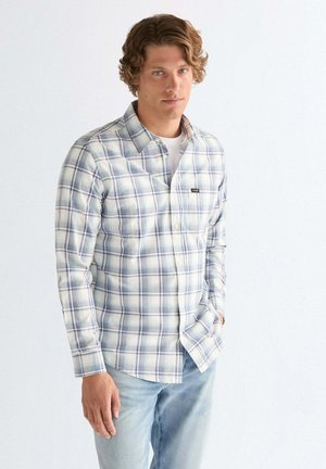 Junger Mann mit lockigem Haar, der ein weiß-blau kariertes Hemd und hellblaue Jeans trägt und mit einer Hand in der Tasche steht.