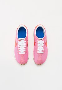 Rosa träningsskor med överdel i mesh och mocka. Har vita snörningar, blå innersulor och ett framträdande orange Nike-swoosh. Gummiyttersula med nubb.