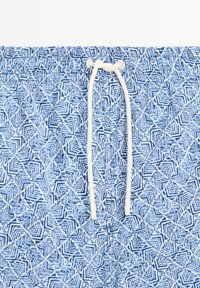 Pantaloni da bagno blu con motivo e strisce geometriche bianche, dotati di una cintura elastica e un cordoncino legato sul davanti.
