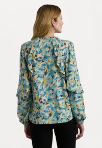 Blusa con stampa floreale in blu chiaro, giallo e marrone. Presenta maniche a volant e un collo arricciato. Tessuto leggero e testurizzato.
