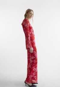 Rød blomstrete lang kjole med gjennomsiktig stoff, lange ermer og en tettsittende silhuett, parret med svarte spisse hæler.