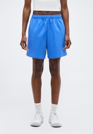 Άτομο που φοράει αθλητικές σορτς με ελαστική μέση σε έντονο μπλε χρώμα, ένα λευκό κοντό τοπ, λευκές κάλτσες και λευκά αθλητικά παπούτσια σε απλό φόντο.