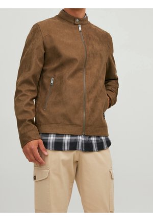 Homme portant une veste en daim marron zippée sur une chemise à carreaux noir et blanc et un pantalon cargo beige, main dans la poche sur fond gris.