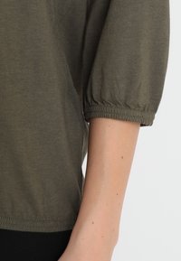 Rechterarm draagt een olijfgroene korte puffmouw shirt met elastische manchetten tegen een witte achtergrond.