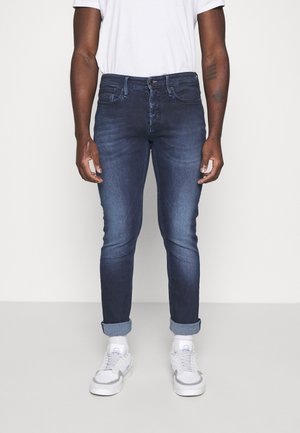 Czarnoskóry mężczyzna w ciemnoniebieskich wąskich jeansach ze zwiniętymi nogawkami, białych trampkach i białej koszulce, stojący na tle gładkiego jasnego tła.