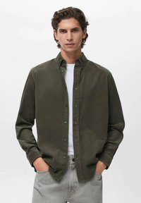 Camisa de botones verde oliva con mangas largas, tela texturada y cuello clásico, llevada sobre una camiseta blanca con pantalones grises.