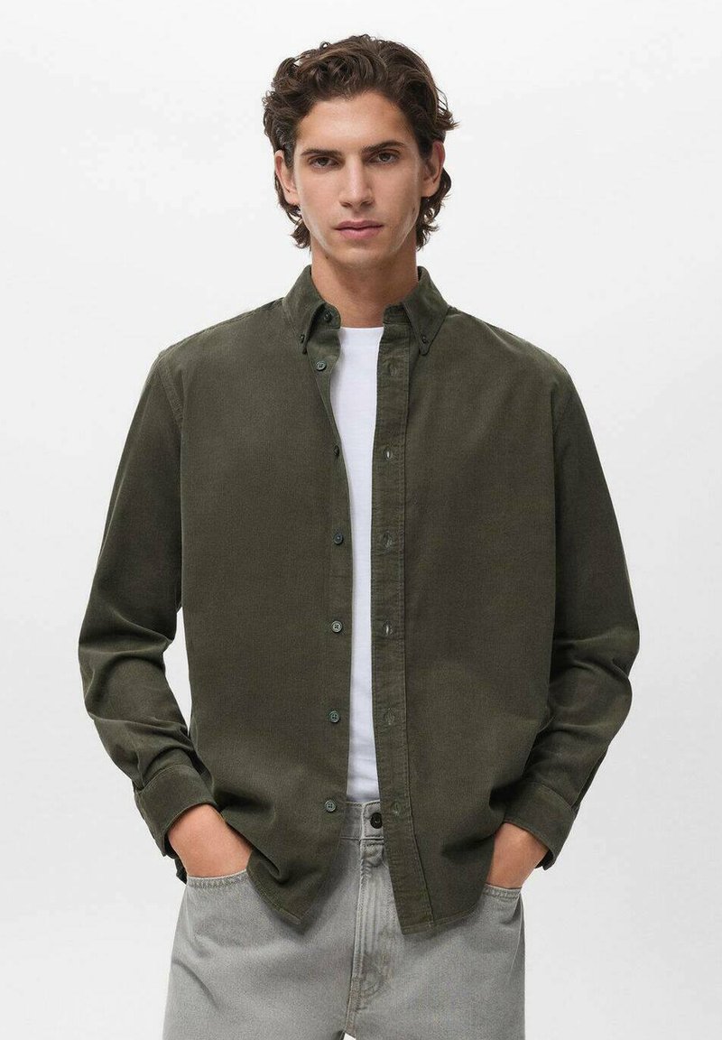 Camisa de botones verde oliva con mangas largas, tela texturada y cuello clásico, llevada sobre una camiseta blanca con pantalones grises.