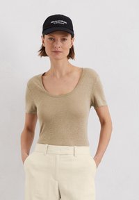 Beige T-shirt met korte mouwen en een ronde halslijn, gemaakt van gestructureerde stof, gecombineerd met crèmekleurige broeken en een zwarte pet met witte letters.