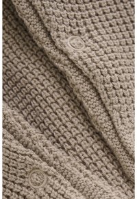 Close-up van beige breiwerk met ronde knoopsluitingen, dat het gestructureerde steeks patroon en gelaagde plooien laat zien.