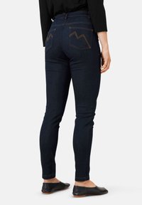 Los jeans skinny de color azul oscuro presentan dos bolsillos traseros con costuras en zigzag naranjas contrastantes. La tela tiene una textura suave, complementando unos zapatos negros.
