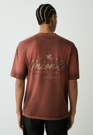 Punutud juustega inimene, kandes tuhmunud pruunikat T-särki, millel on tekst "Union Co Montana Great Falls" ja seljal väike tiivuline kujund.