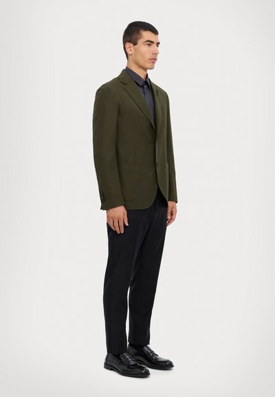 Sacou verde măsliniu cu un singur nasture, rever clasic și două buzunare frontale, purtat cu pantaloni negri și pantofi negri lucioși.
